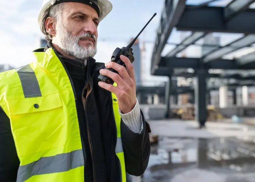 A professional construction supervisor wearing a white safety helmet and high-visibility vest using a handheld two-way radio for real-time team coordination on a building site